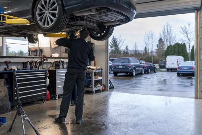 A tidy service bay where a mechanic, back to the camera, works beneath a car lifted on a hoist