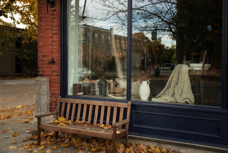 An independent boutique window set in a brick wall in autumn, with a wooden bench out front
