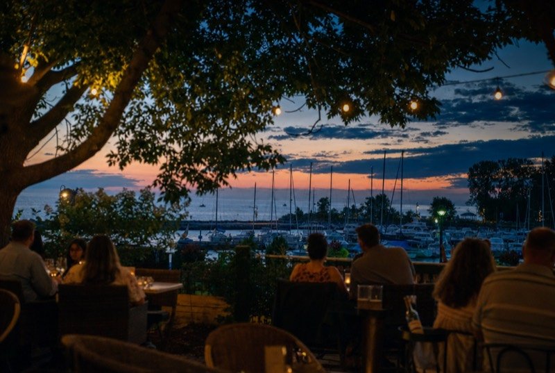 String lights over a restaurant patio at dusk, looking out on a small harbour of moored sailboats
